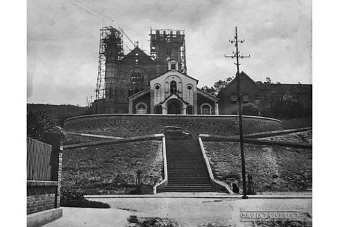 Foto em preto e branco, tirada a partir da Avenida dos Andradas. Há uma marca d'água no canto inferior direito da imagem, com os escritos “www.MariadoResguardo.com.br”. A imagem apresenta, em primeiro plano, uma vista frontal da escadaria em frente à Capela da Glória. Ao fundo, logo atrás da Capela, é possível visualizar a Igreja da Glória ainda em construção. À direita da Igreja, há uma outra edificação. A escadaria é larga e começa a partir da Avenida dos Andradas e termina no Largo Da Glória, rua abaixo da Capela. No topo do morro, a fachada frontal da Capela é ampla, possui janelas arqueadas nas laterais e, ao centro, uma grande entrada também arqueada. Ao fundo, é possível visualizar diversos andaimes envolvendo a Igreja da Glória. A Igreja é ampla, alta e dispõe de um volume central com cobertura em dois caimentos com uma cruz no topo. Há duas torres laterais ainda em construção.