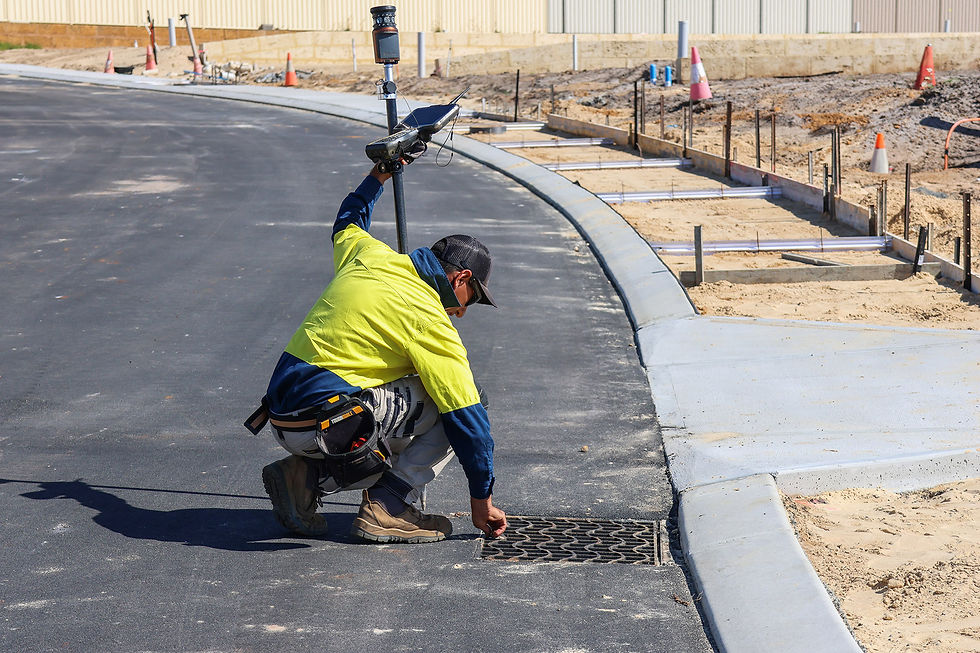 Construction worker crouches by a storm drain on a new asphalt road at a sandy building site, checking the grate.
