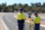 Two men stand on a road, setting up surveying technology. They both wear yellow hi-vis shirts.