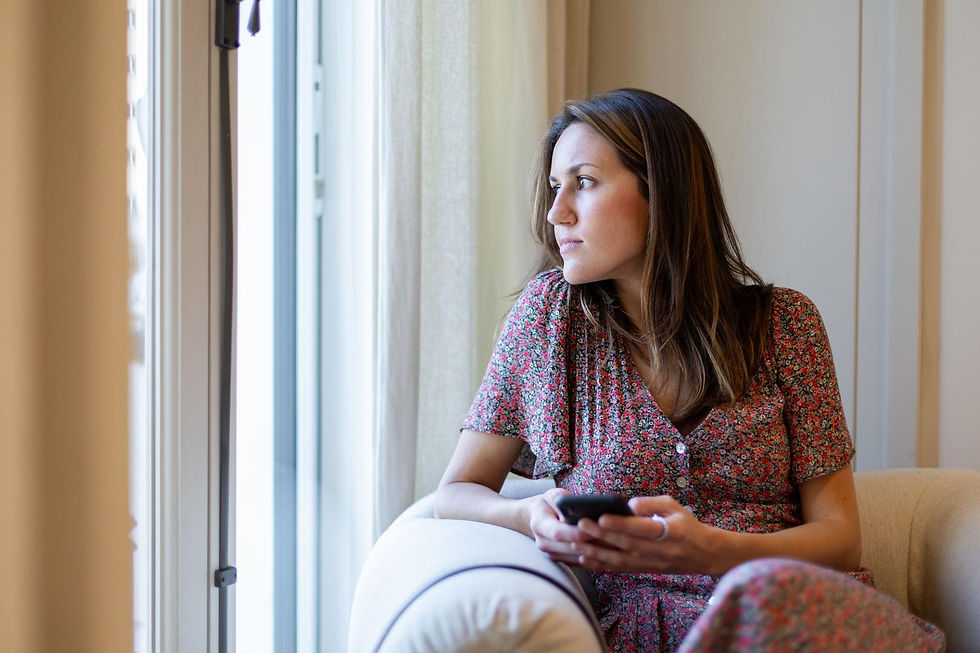 Une femme adulte assise près d'une fenêtre, un téléphone à la main avec une expression pensive. Cette scène évoque la peur de l'abandon, l'inquiétude face à un message sans réponse et le besoin de réassurance dans les relations affectives