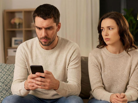 Deux adultes en couple sont assis sur un canapé, dans un atmosphère tendue. L'un tient un téléphone tandis que l'autre semble préoccupé. La scène évoque les difficultés de confiance, les demandes de réassurance et le contrôle dans le couple