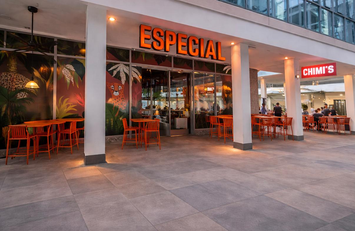 Mostly empty outdoor tables and orange chairs outside restaurants in the early evening