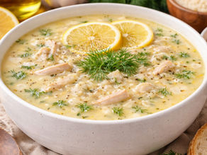 Creamy soup with chicken, rice, dill, and lemon slices in a white bowl on a wooden table. Bread slices and lemon wedges nearby.