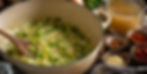 Chopped green peppers and onions are being cooked in a cream pot with a wooden spoon. Surrounding ingredients include spices and broth.