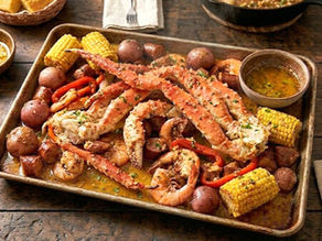 Seafood boil on a tray with crab legs, shrimp, corn, and potatoes. Bowls of butter sauce, cornbread, lemon wedges. Rustic wood table.