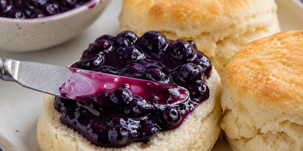 Biscuits with blueberry jam being spread by a knife. The jam is dark purple and glossy. A bowl of jam is visible in the background.