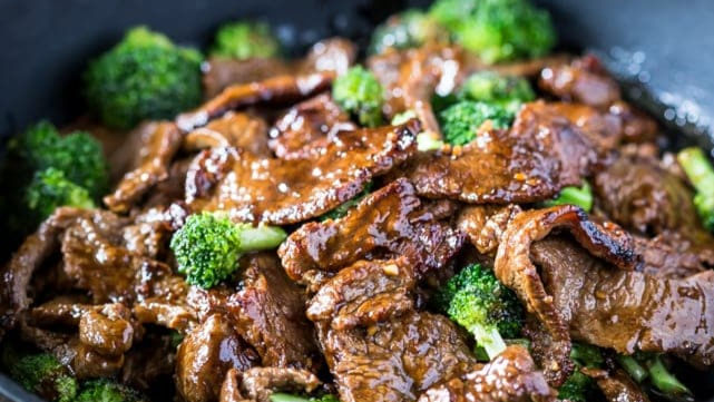 Close-up of beef and broccoli stir-fry, glistening with sauce. Vibrant green broccoli contrasts with the rich brown beef in a pan.