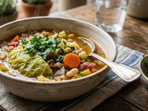 A bowl of vegetable soup with parsley on a wooden table, next to toasted bread and rosemary. Potted plants in the background.