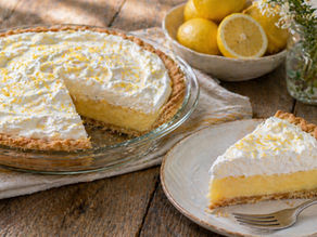 Lemon pie with whipped cream and zest, slice on plate with fork. Lemons and daisies in background on rustic wooden table.