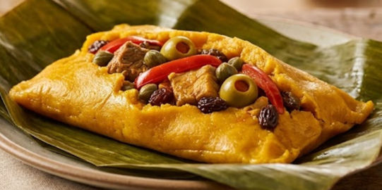 Tamale on a green leaf, topped with olives, peppers, meat, and raisins. Bright colors, rustic wooden table in the background.