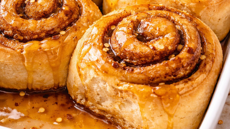 Close-up of cinnamon rolls drizzled with caramel sauce, with golden-brown swirls and glossy surface, in a white baking dish.