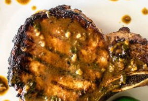 Grilled pork chop with a golden-brown crust on a white plate drizzled with sauce, creating a savory and appetizing presentation.