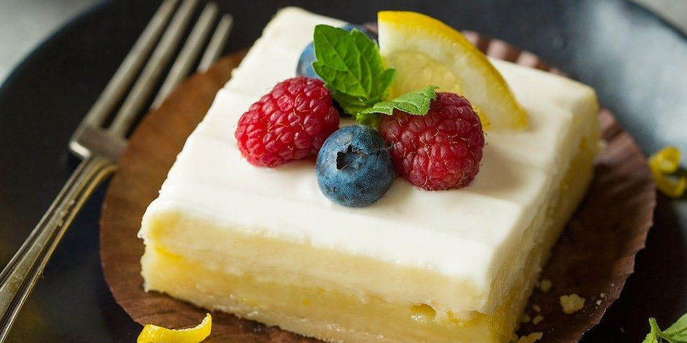 A square lemon dessert topped with blueberries, raspberries, a lemon slice, and mint on a dark plate with a fork on the side.