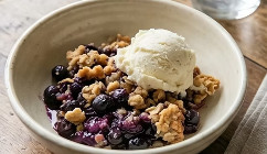 Warm blueberry crisp topped with a scoop of vanilla ice cream and crunchy oat crumble, served in a rustic bowl with juicy blueberries visible underneath.