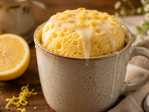 Lemon mug cake topped with glaze in a speckled mug, with whole and sliced lemons on a rustic table and a spoon nearby