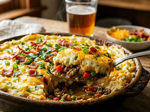 A casserole with cheese, bacon, and herbs is being served in a cozy kitchen. A glass of beer and a bowl of toppings are in the background.