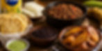 Venezuelan meal with shredded beef in a pot, grilled arepas, plantains, black beans, cheese, green sauce, and a maize flour pack on a wooden table.