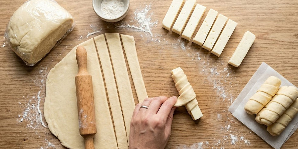 A hand rolls dough strips with cheese on a wooden table. Dough, flour, rolling pin, and wrapped pastries nearby convey a baking scene.