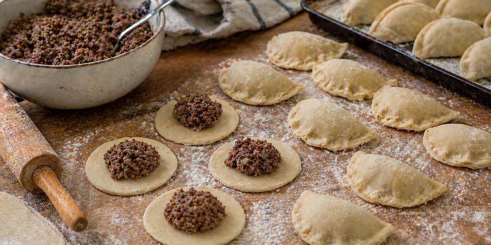 Dough circles filled with minced meat on a floured wooden table. A rolling pin and a bowl of filling are visible, ready for assembly.