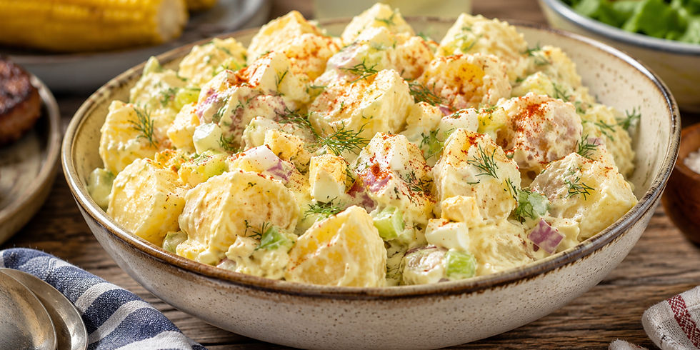 Bowl of creamy potato salad with dill, sprinkled with paprika. Corn, steak, and green salad visible in the rustic wooden background.