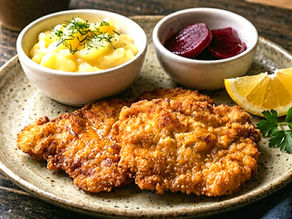 Crispy schnitzel with lemon wedge on plate. Served with mashed potatoes topped with dill and sliced beets. Rustic wooden table setting.