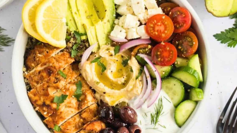 A colorful bowl with grilled chicken, avocado, hummus, cherry tomatoes, cucumbers, red onion, olives, and lemon slices on a white surface.