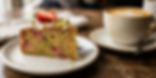 Slice of cake with icing and pistachios on a plate, beside a halved strawberry. Cup of coffee in the background. Warm, inviting mood.