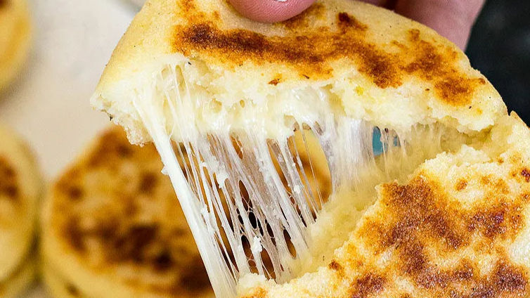 Close-up of a hand pulling apart a cheesy arepa, showing melted cheese stretching. Golden brown crust, background of multiple arepas.