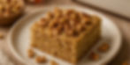 Square slice of cake topped with walnuts on a white plate. Wooden table and a bowl of walnuts in the background. Warm, inviting mood.