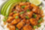 Bowl of glazed chicken pieces with sesame seeds and green onions, sliced avocado, lime wedge, and rice. Vivid colors, appetizing look.