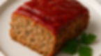 A slice of meatloaf topped with glossy red sauce on a white plate, garnished with fresh parsley. Neutral background.