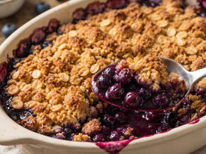 Close-up of a blueberry crumble in a white dish with a spoon scooping a portion. Golden crust, vibrant purple filling, blueberries scattered.