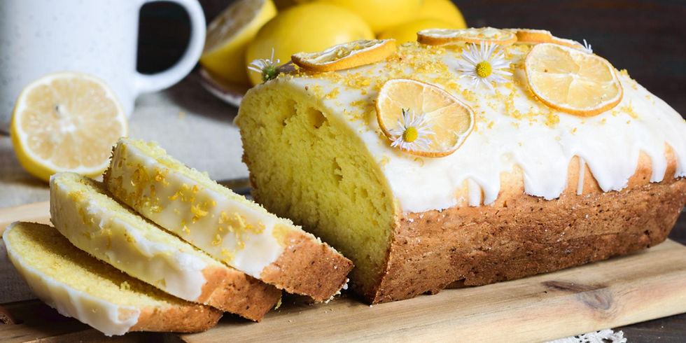 Lemon cake with white icing, topped with lemon slices and flowers, sits on a wooden board with lemons and a mug in the background.
