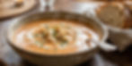 Creamy tomato soup with herbs and cream swirl in a rustic bowl on wooden table. Steamy and warm, with sliced bread in the background.