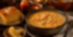 A bowl of creamy soup with crab, parsley, cornbread on the side, lemon slices in a dish, and hot sauce in a rustic setting.