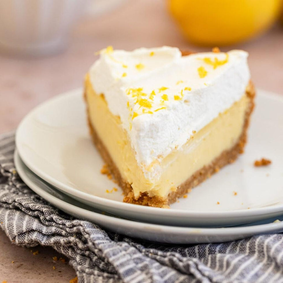 Slice of lemon pie with whipped cream, lemon wedge, and mint on a white plate. Crumbly crust and blue background evoke a fresh mood.