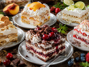 Five plated slices of homemade icebox cake arranged on a rustic farmhouse table, including banana pudding, strawberry, chocolate cherry cheesecake, peach, and key lime flavors. Fresh fruit, graham crackers, chocolate pieces, citrus, and mint surround the desserts, while full glass baking dishes of each icebox cake sit softly blurred in the background under warm natural light.