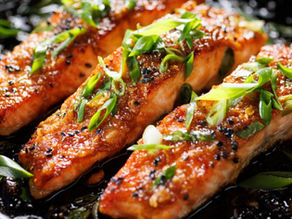 Three glazed salmon fillets topped with green onions and black sesame seeds in a dark skillet. The setting is vibrant and appetizing.