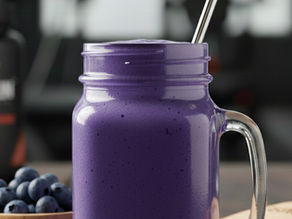 Purple smoothie in a mason jar with straw, on a wooden table. Bowl of blueberries nearby, protein powder jar, and water bottle in background.
