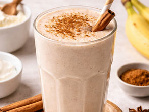 Creamy smoothie in a clear glass, topped with cinnamon and a straw. Surrounded by bananas, cinnamon sticks, and white bowls on a light surface.