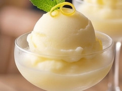 A scoop of smooth lemon sorbet served in a glass dessert dish, garnished with lemon zest and fresh mint in a bright summer setting.