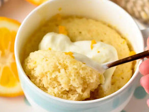 A spoon scoops lemon mug cake with cream from a white cup. Orange slices and striped cloth in background. Hands with red nail polish.