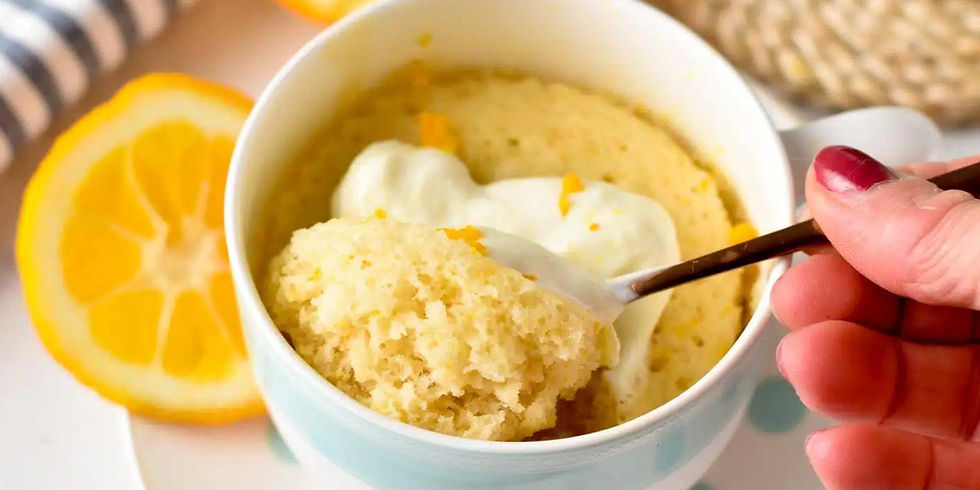 A spoon scoops lemon mug cake with cream from a white cup. Orange slices and striped cloth in background. Hands with red nail polish.