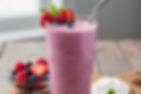Pink smoothie in a tall glass with a metal straw, garnished with berries and mint. Bowl of berries and whipped cream on wood table.