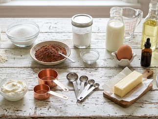 Ingredients for Red Velvet Cupcakes, including flour, cocoa powder, buttermilk, butter, egg, vanilla, red food coloring, and baking essentials arranged on a rustic countertop.