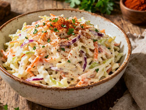 Bowl of creamy coleslaw with carrots and red onions, topped with paprika and parsley, on a rustic wooden table with a cloth and spices.