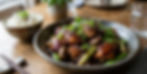 Bowl of glazed chicken with green onions and sesame seeds, next to a bowl of rice with black sesame seeds on a wooden table. Cozy setting.