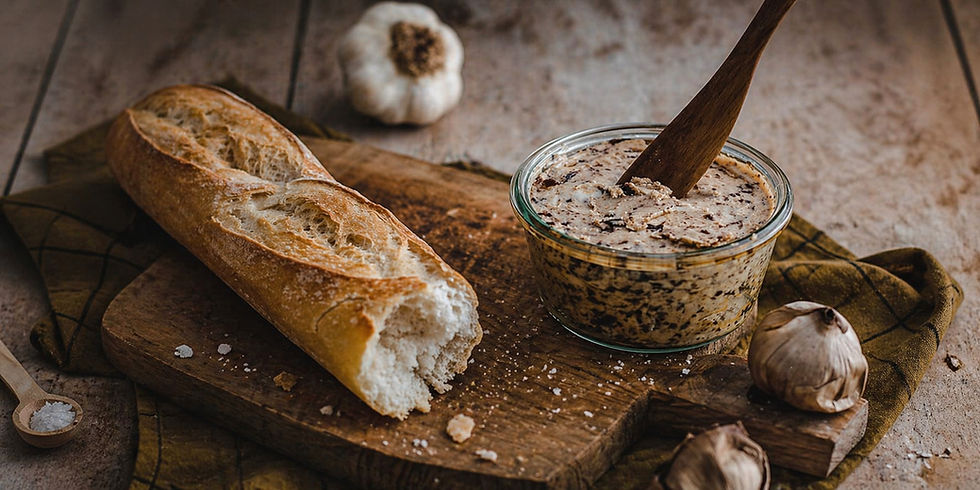 Baguette and glass jar of spread on a wooden board, surrounded by garlic cloves and a wooden spoon, on a rustic table.