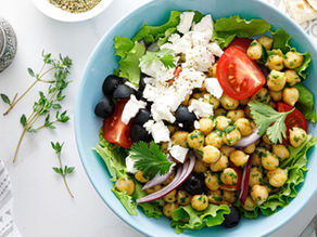 Chickpea salad with tomatoes, feta, olives, and greens in a blue bowl. Silver grinder, herbs, cloth, and pita on a white table.