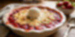 Fresh strawberry cobbler in a pie dish topped with vanilla ice cream, beside a bowl of strawberries and a spoon on a rustic table